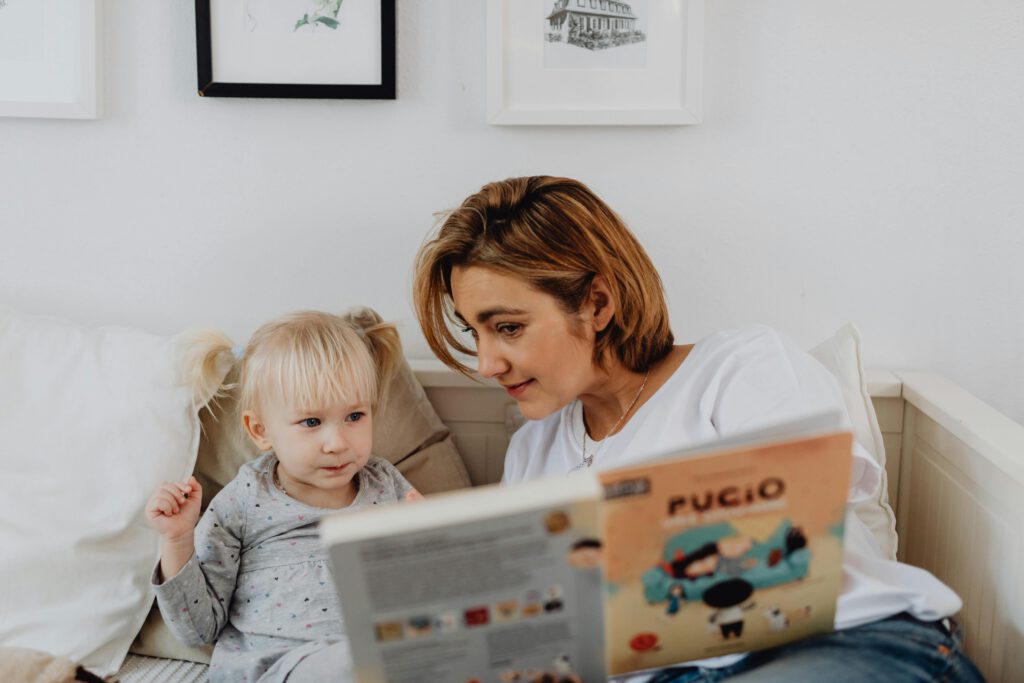 A mother and her daughter sharing a bonding moment while reading a children's book in the bedroom.